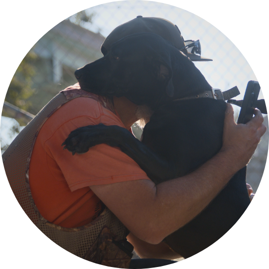 Black dog standing on legs with paws up on Paul's shoulders to give him a hug as he's crouched down.